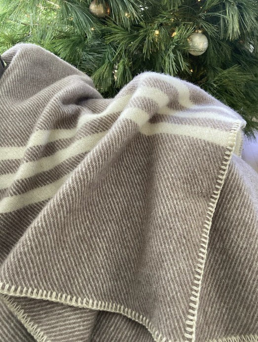 A natural taupe wool throw with natural stripes, draped over a surface with a Christmas tree in the background.