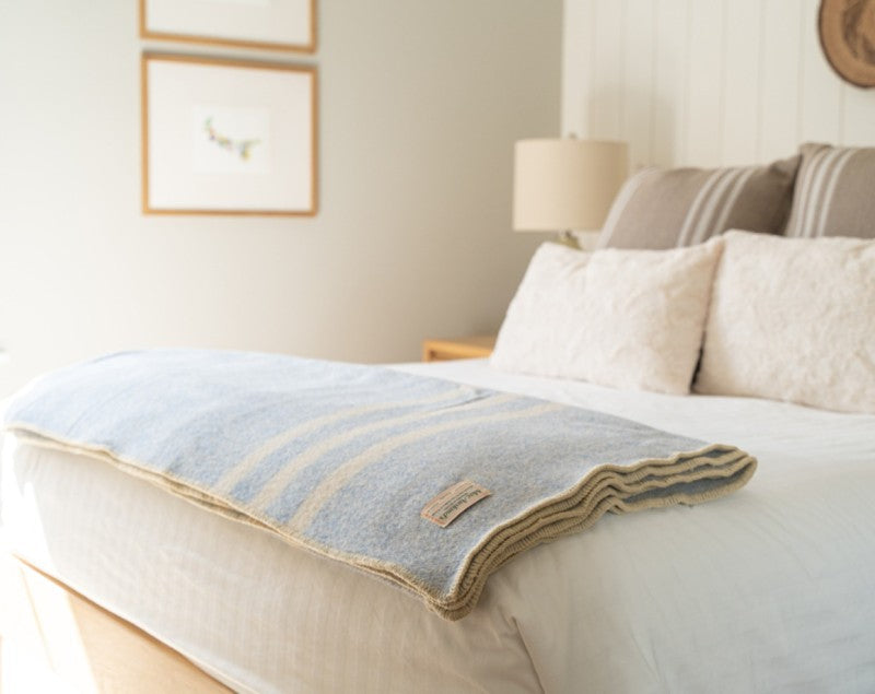 A throw blanket in blue tweed with white wool throw draped over the end of a bed, with pillows and a lamp in the background.