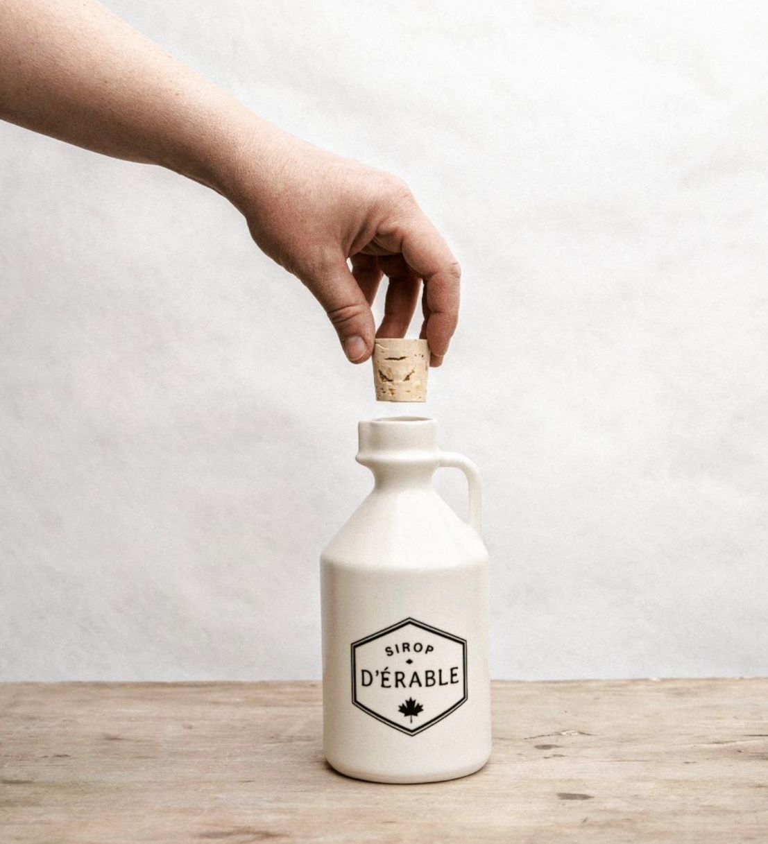 A person's hand placing a cork on a white ceramic maple jug with a label that reads 'SIROP D'ERABLE' and has a small decorative Canadian Maple leaf on it